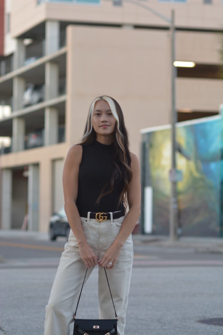 Realtor standing on a sidewalk with one leg forward wearing a black top and beige pants