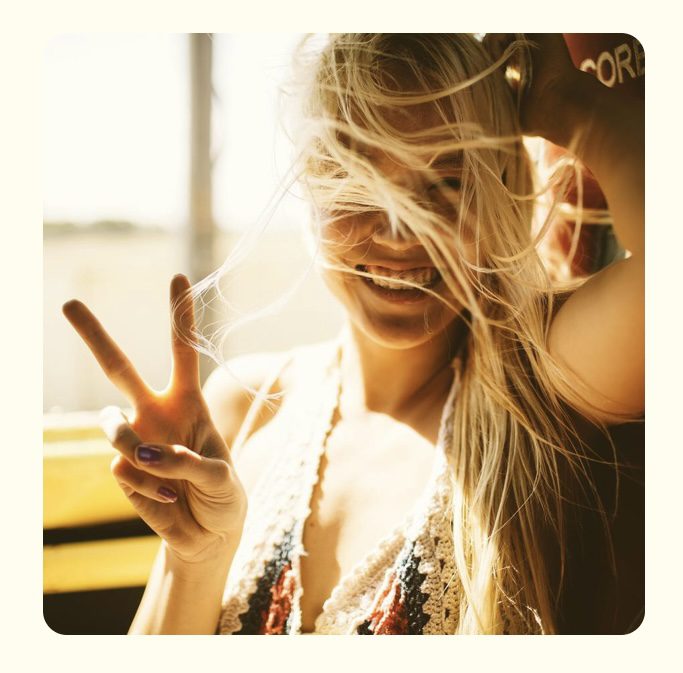 Blonde woman in her 30s laughs in a sunlit window throwing a peace sign in a crochet top