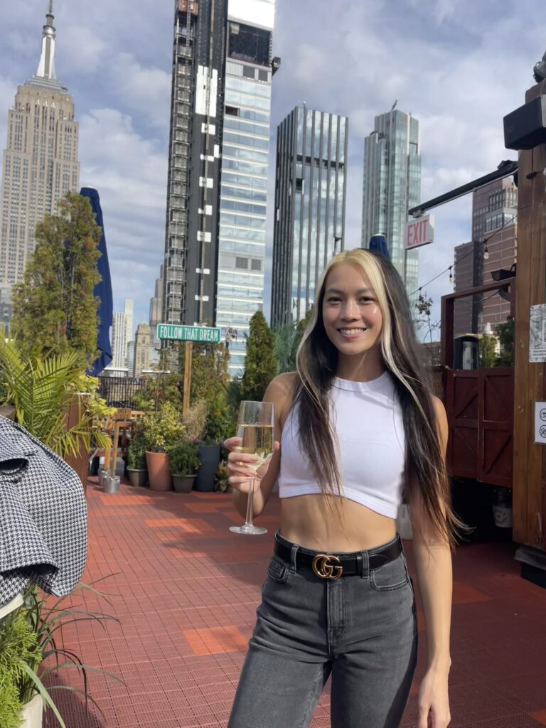 Smiling woman in her 30s holds champagne on a rooftop overlooking the New York City skyline
