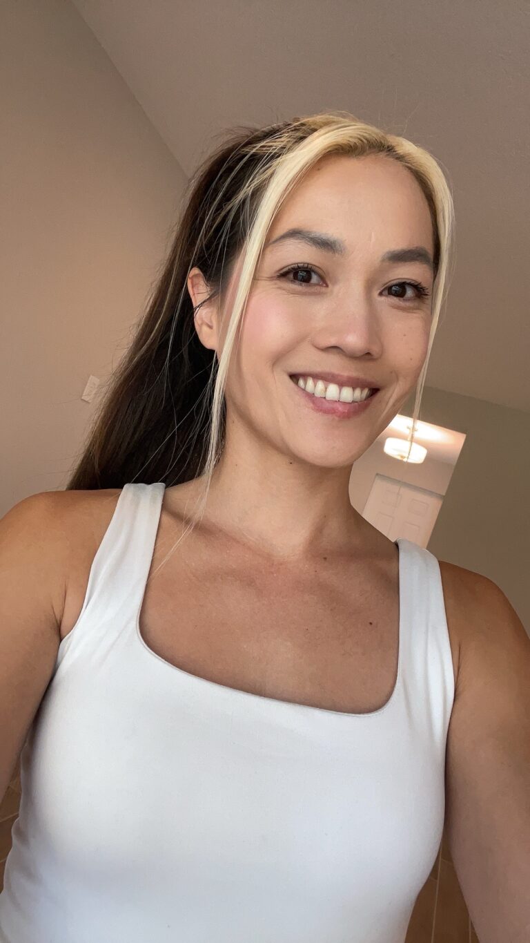 Woman in her 30s smiling in white tank top taking a natural no-makeup skincare selfie indoors