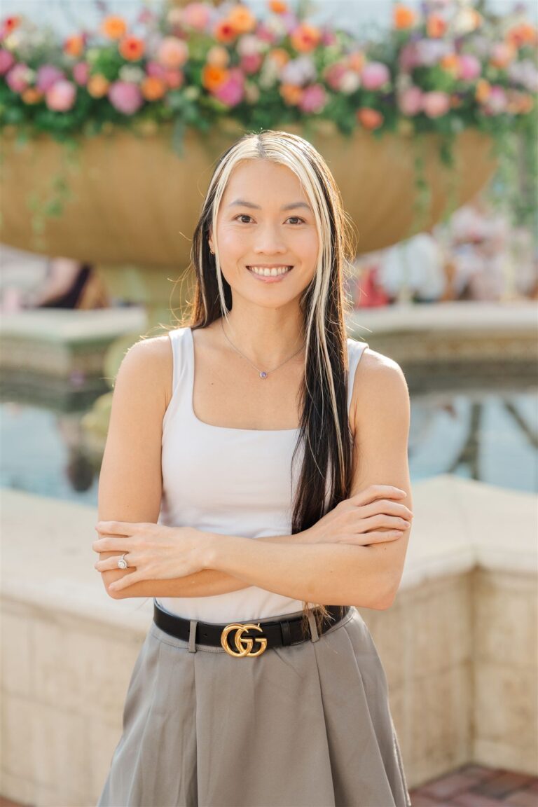 Smiling woman in her 30s in white tank top and gray skirt by a flower-topped fountain