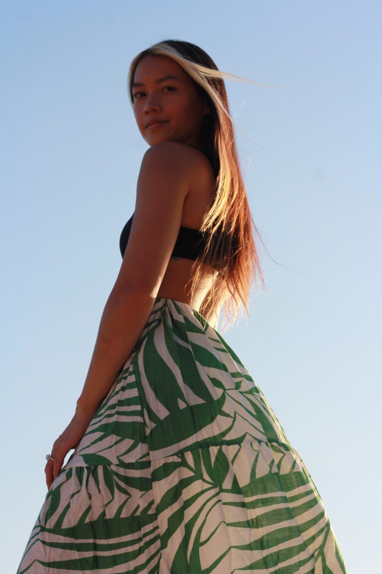 Woman in her 30s in black top and green palm-print maxi skirt against blue sky at sunset