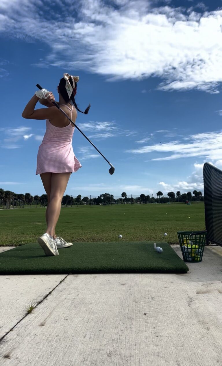 Lizzie at the top of her golf swing at a driving range in a pink gingham golf dress
