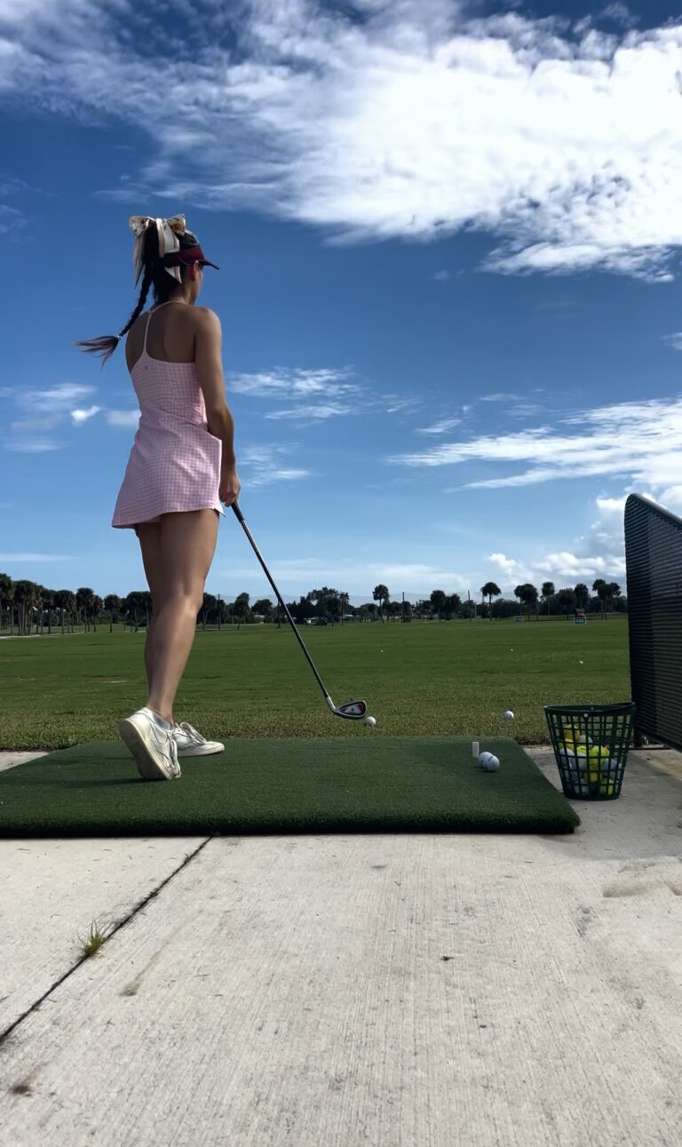 Lizzie at a driving range in a pink gingham golf dress, visor with a bow and white sneakers, set up to hit a ball